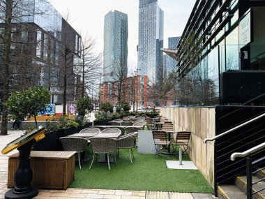 Manchester, İngiltere - 24 Ocak 2026: Manchester 'daki Gino D' Acampo cafe alanında hasır sandalyeler, beton ekiciler ve uzun cam kuleler.