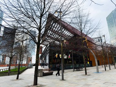 Manchester, İngiltere - 24 Ocak 2026: Manchester 'da cam-önlü Hint Tiffin Odası, yapraksız ağaçlar ve dekoratif ışıkların yer aldığı çağdaş bir sokak manzarası.