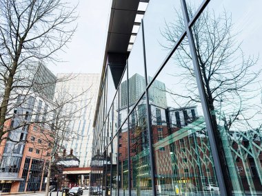 Manchester, İngiltere - 24 Ocak 2026: Modern cam binalar kışın çıplak ağaçlar ve kentsel mimari ile Manchester caddesini yansıtıyor.
