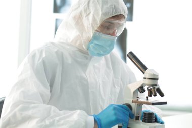 Laboratuvarda mavi eldiven takan koruyucu kıyafetli bir doktor tıbbi mikroskopla bakar.