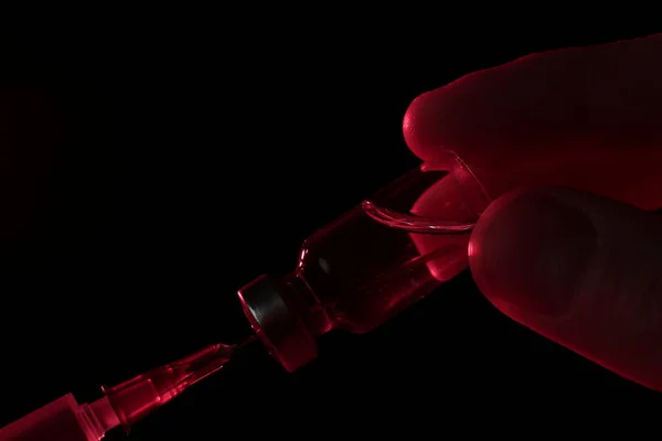 Doktorun elleri kırmızı ışıkta siyah arka plandaki cam ampulden şırıngaya aşı yazıyor.
