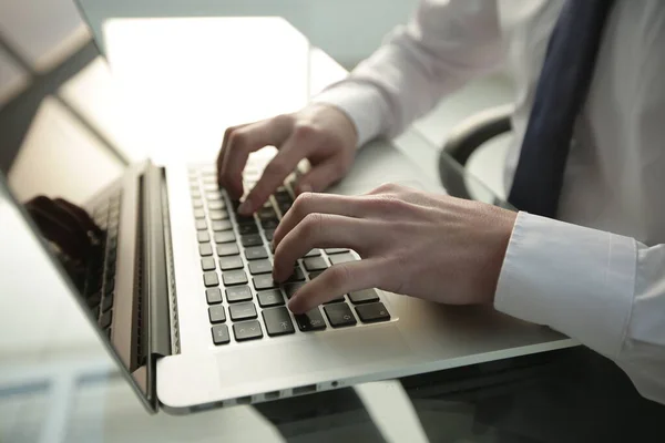 Beyaz gömlekli ve kravatlı bir yönetici cam masaya oturur ve geniş parlak bir ofiste dizüstü bilgisayara yazar.