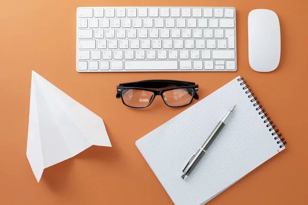 Brille weiße Tastatur-Maus-Notizblock und Papierflieger auf orangefarbenem Hintergrund — Stockfoto