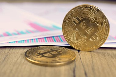 Bitcoin kripto parasının bir altın sikkesi kendi ucunda duruyor, ikincisi ise ofis masasındaki ahşap bir masada kripto para değerlerindeki değişimlerin çizelgesinde yatıyor.