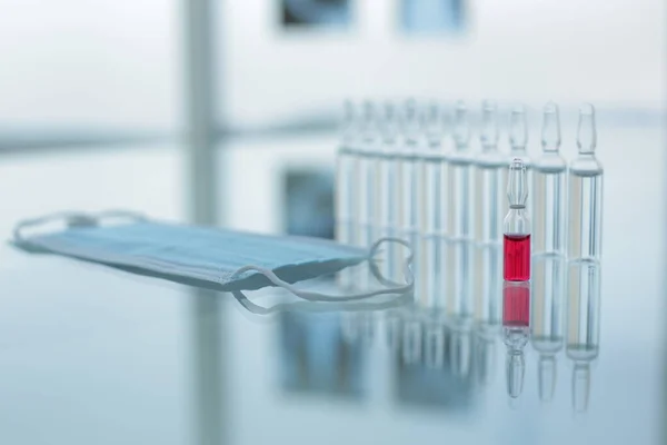 Vial con vacunas rojas y transparentes máscara de protección médica en la mesa de vidrio en la clínica —  Fotos de Stock