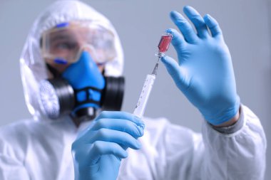Koruyucu takım elbiseli bir doktor solunum cihazıyla mavi eldivenli bir şırıngadan aşı çeker.
