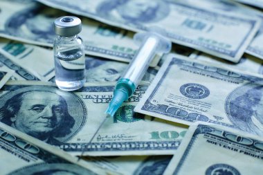 100 dolarlık banknotların arka planında aşı şişesi ve şırınga var.