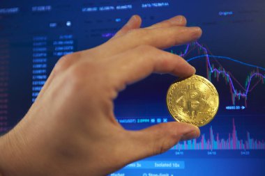 Bir adam elinde altın bir bitcoin tutuyor. Şifreleme kurlarındaki değişikliklerin grafikleri dizüstü bilgisayar monitöründe gösterilir