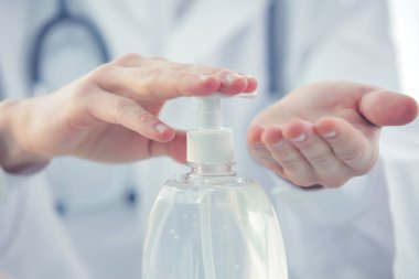 Klinikte steteskoplu ve koruyucu maskeli bir doktor ellerini antiseptikle tedavi ediyor. Yakın plan.
