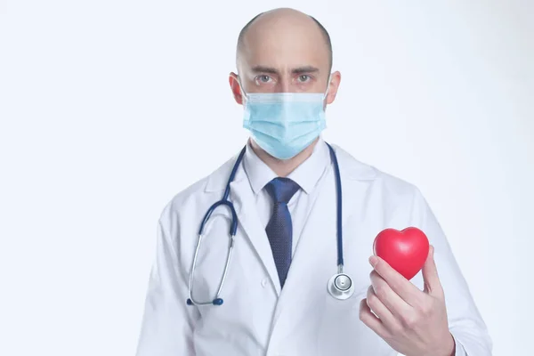 Doctor en máscara protectora y estetoscopio médico sostienen el corazón en su mano. Aislado en fondo blanco — Foto de Stock