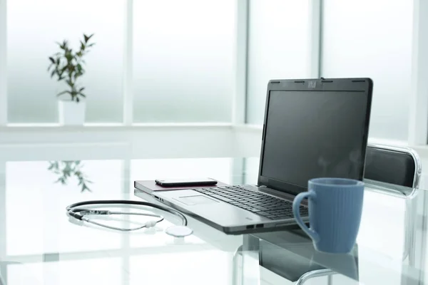 Laptop medikal steteskop kahve fincanı telefon günlüğü planlayıcısı doktorların parlak ofis alanındaki cam masanın üzerinde.