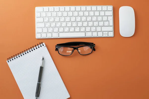 Glasses mouse keyboard notepad and pen on orange background