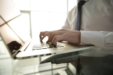 Beyaz gömlekli ve kravatlı bir yönetici cam masaya oturur ve geniş parlak bir ofiste dizüstü bilgisayara yazar.