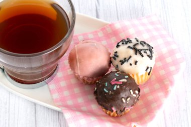 Bir fincan çay, çeşitli tat ve renklerde küçük kekler eşliğinde. Pembe ve beyaz ekose peçeteyle yerleştirilmiş tepsi..