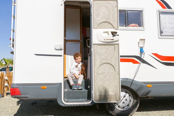 Küçük kıvırcık saçlı çocuk karavanın kapısında oturmuş gülümsüyor, şefkatle evcil hayvanını tutuyor..