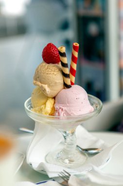 Üç çeşnili dondurma, yanında renkli spiral gofretler, hepsi güzel cam bir fincanda sunuluyor, üstünde çilek var..