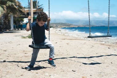 Kıvırcık saçlı beyaz çocuk, kumlu sahilde denizin kenarında bir salıncakla oyun alanında eğleniyor..