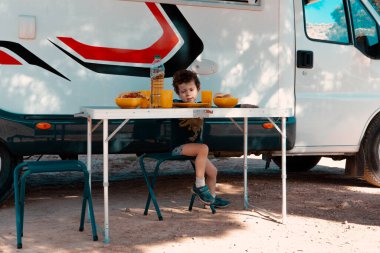 Bir ağacın gölgesinde kamp koltuğunda oturan küçük bir çocuk masadaki yemeği yemeye hazır..