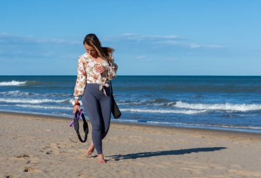 Çıplak ayaklı kadın sahilde yoga minderi ve fitness yüzüğü taşıyarak yürüyor. Okyanus kenarında güneşli bir günün tadını çıkarıyor..
