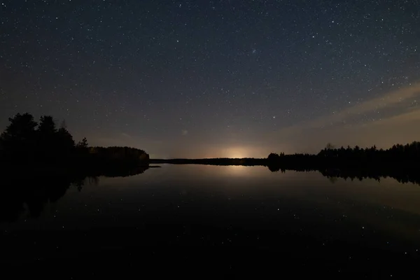 Yıldızlı gece gökyüzü sessiz bir orman gölü yüzeyine yansıyor
