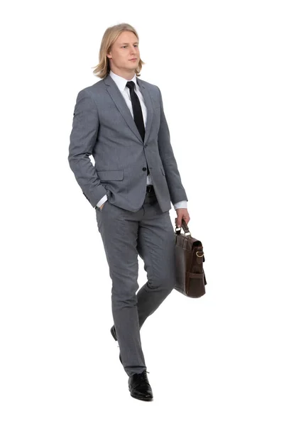 Gri takım elbiseli genç bir işadamı beyaz arka planda izole edilmiş bir çanta taşıyor ve yürüyor.