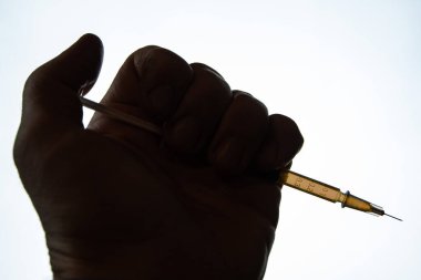 Bağımlının koyu renkli eli, parlak ışığın arka planına karşı içinde uyuşturucu çözeltisi bulunan bir şırınga tutar.