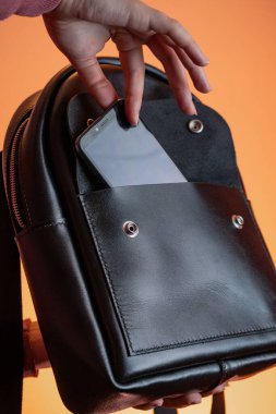 Bir kadının eli, turuncu bir arka plandaki deri bir sırt çantasının cebinden telefonu çıkarır.