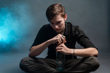 Depresyonda olan genç bir adam, elinde bir şişeyle, gri bir arka planda oturur.