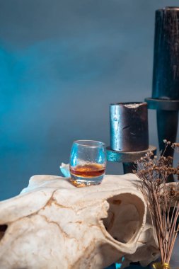 Kafatasının üzerinde bir bardak rom duruyor. Arka planda siyah mumlar, gri bir arka planda.