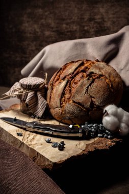 Bıçak bir tahta kütüğün üzerinde, bir ekmeğin yanında ve koyu bir arka planda bir kavanoz kuru duruyor.