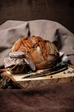 Bıçak bir tahta kütüğün üzerinde, bir ekmeğin yanında ve koyu bir arka planda bir kavanoz kuru duruyor.