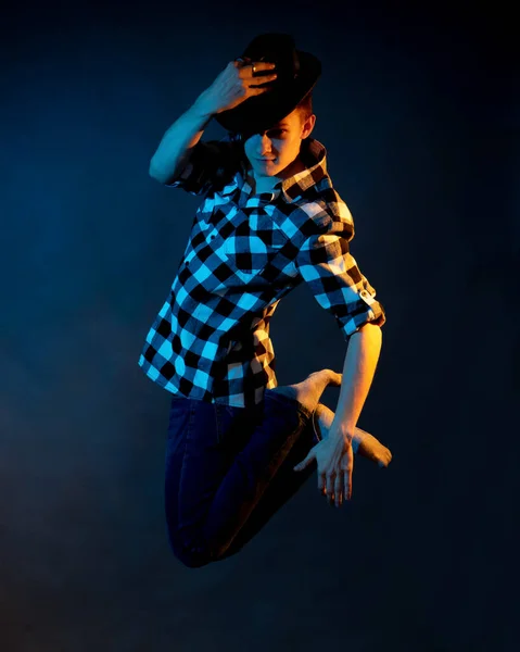 Un gars en chemise à carreaux et chapeau saute sur un fond sombre avec une lumière bleue et jaune — Photo