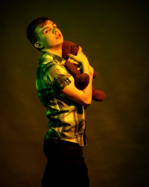 Genç bir adam, sarı ve yeşil ışıkla aydınlatılmış bir oyuncak ayıya sarılıyor.