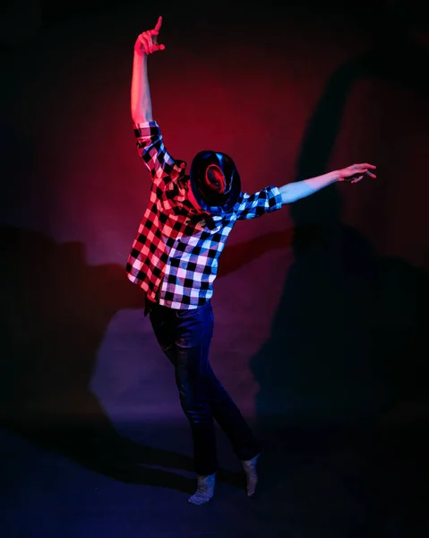 Un jeune homme en chemise à carreaux et chapeau en cuir danse, illuminé par la lumière rouge et bleue sur un fond sombre dans le studio — Photo