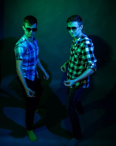 Two young men in plaid shirts dance in the studio with multicolored light — Stock Photo, Image