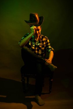 Ekose gömlekli, kovboy şapkalı genç bir adam yeşil ve sarı ışıkla aydınlatılmış bir sandalyeye oturur.