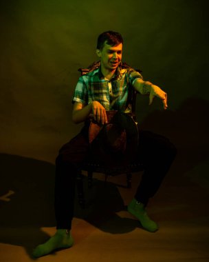 Ekose gömlekli, kovboy şapkalı genç bir adam yeşil ve sarı ışıkla aydınlatılmış bir sandalyeye oturur.