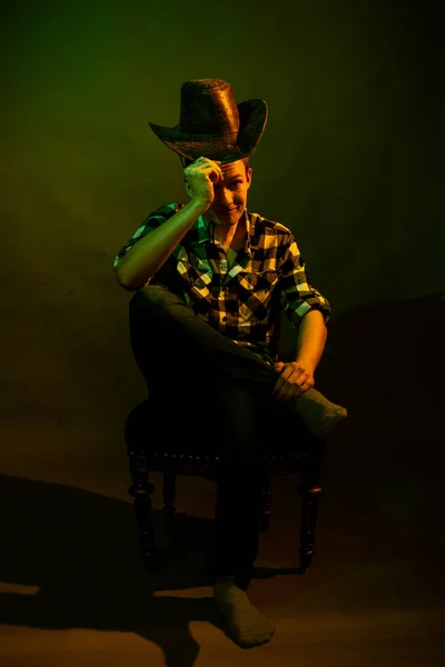 En ung man i rutig skjorta med cowboyhatt sitter på en stol, upplyst av ett grönt och gult ljus — Stockfoto
