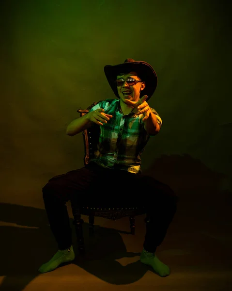 Un jeune homme en chemise à carreaux avec un chapeau de cow-boy assis sur une chaise, illuminé par une lumière verte et jaune — Photo