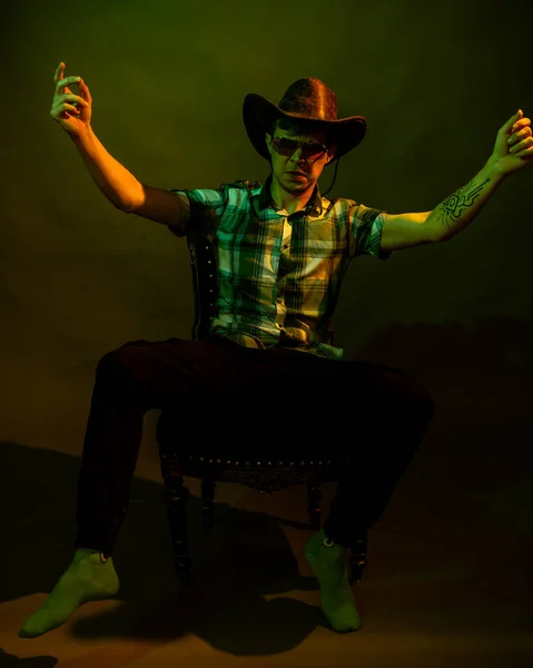 Un joven con camisa a cuadros con sombrero de vaquero se sienta en una silla, iluminado por una luz verde y amarilla — Foto de Stock