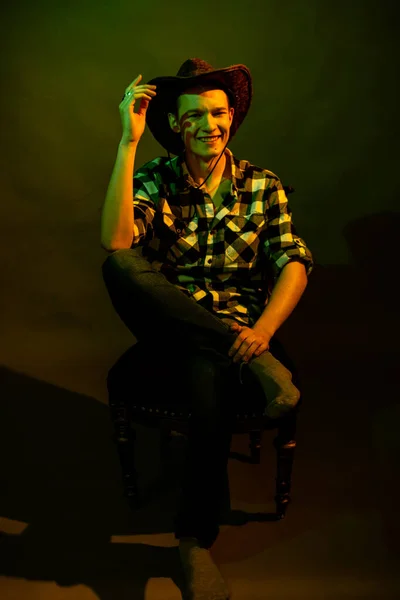 Un jeune homme en chemise à carreaux avec un chapeau de cow-boy assis sur une chaise, illuminé par une lumière verte et jaune — Photo