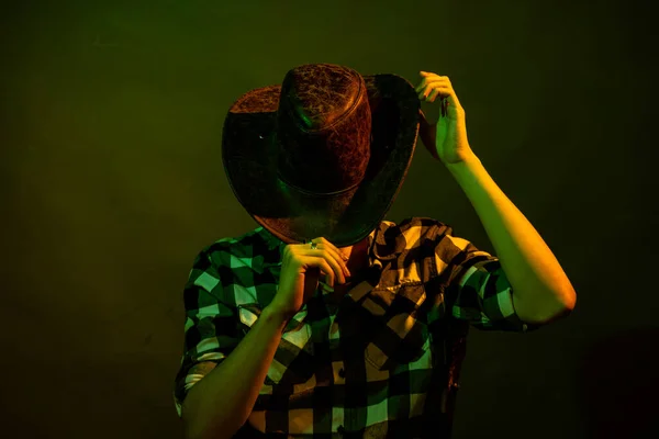 Un jeune homme en chemise à carreaux avec un chapeau de cow-boy assis sur une chaise, illuminé par une lumière verte et jaune — Photo