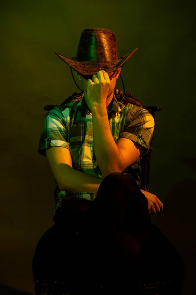 Un joven con camisa a cuadros con sombrero de vaquero se sienta en una silla, iluminado por una luz verde y amarilla — Foto de Stock