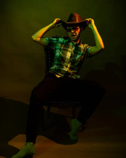 一个穿着格子呢衬衫、头戴牛仔帽的年轻人坐在椅子上，绿色和黄色的灯光照亮着他 — 图库照片