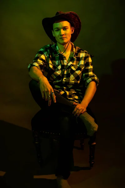 Молодой человек в клетчатой рубашке в ковбойской шляпе сидит на стуле, освещенном зеленым и желтым светом — стоковое фото