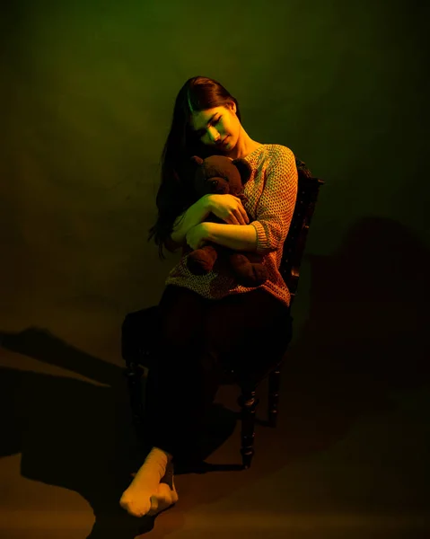 A cute girl with a teddy bear sits on a chair in the studio, illuminated by yellow and green light — Stock Photo, Image