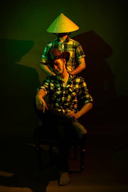 Kovboy ve Çin şapkalı iki adam koyu bir arka planda sarı ve yeşil ışıkla aydınlatılmış.