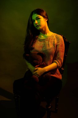 Stüdyoda elinde oyuncak ayıyla oturan sevimli bir kız, sarı ve yeşil ışıkla aydınlatılmış.