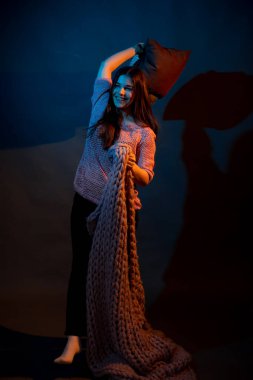 Pembe kazaklı bir kız, sarı ve mavi ışıkla aydınlatılmış yastıklar tutuyor.