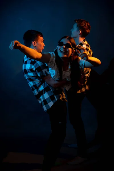 Två killar i rutig skjorta håller en flicka på en mörk bakgrund i en studio med flerfärgat ljus — Stockfoto